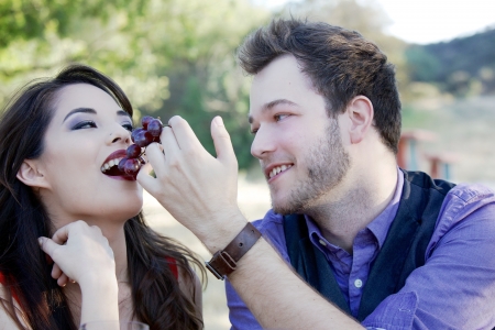 A young 20-something man feeds a 20-something woman grapes from the vine, while in a park picnicking