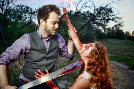 A mans is about to behead a grotesque and bloody female zombie in a wooded area