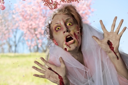 Grotesque and bloody female zombie in a wedding dress and veil stands in a field of spring time cherry blossom treesの写真素材