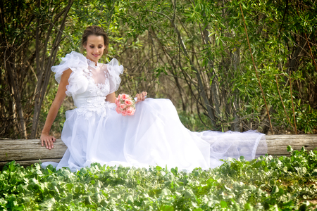 A smiling young bride dressed in a flowery and puffy white wedding dress sits on a wooden log in a grove of trees  の写真素材