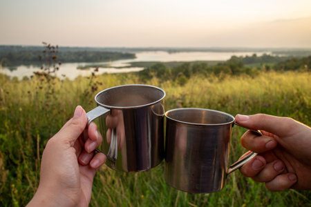 Two hands holding stainless steel camping mugs in a toast against a scenic sunset landscape with rolling hills and a winding river symbolizing outdoor adventure travel and shared moments in nature.の写真素材