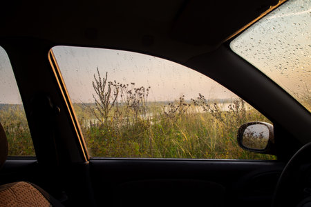Looking out from inside a car through a window covered in raindrops reveals a hazy landscape of vegetation and a body of water under a soft evening light suggesting a journey on a wet road.の写真素材
