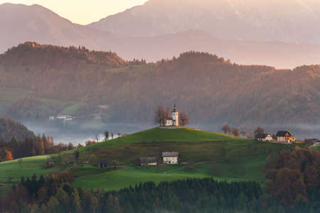St. Thomas Church with the low lying fog and the alps in the background during the sunrise of the fall season of Sloveniaの写真素材