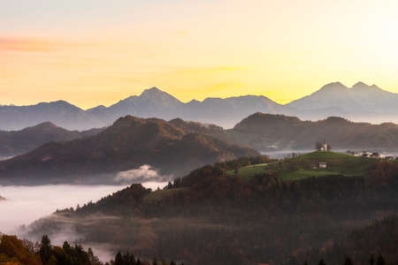 Skofja Loka, Slovenia - view of the beautiful Church of St. Tomaz St. Thomas on top of the hill with an amazing misty sunrise and the Julian Alps in the background in the fall.の写真素材
