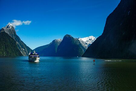 A tourist sightseeing ferry boat travels down the calm, deep blue waters of Milford Sound in the remote Fiordland region in the south west of the South Island of New Zealandの写真素材
