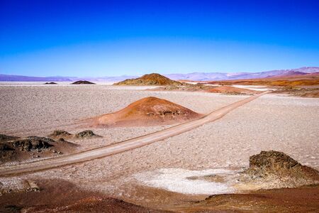 Red 4wd tracks cut across a salt flat punctuated by red and orange hills, in the high altitude altiplano puna desert near Salta in Argentinaの写真素材