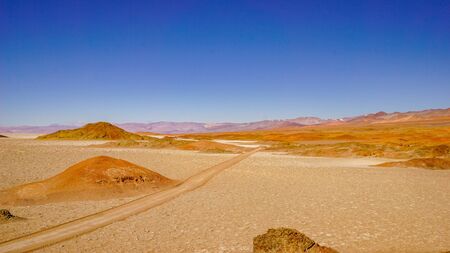 Red 4wd tracks cut across a salt flat punctuated by red and orange hills, in the high altitude altiplano puna desert near Salta in Argentinaの写真素材