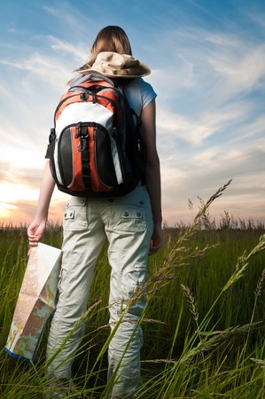 beautiful young woman with backpack and map in hand standing outside in the field with her back to...