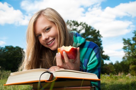 beautiful young student girl lying on grass with red apple in her hand and books under her hands, looking into the camera and smiling. Blue cloudy sky in backgroundの写真素材