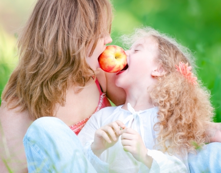 Beautiful and  happy young mother eating apple together with her small daughter. Summer park in backgroundの写真素材
