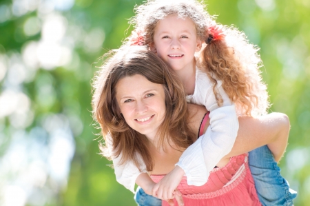 Beautiful and happy young mother giving piggyback ride to her daughter. Both smiling and looking into the camera. Summer park in background.の写真素材