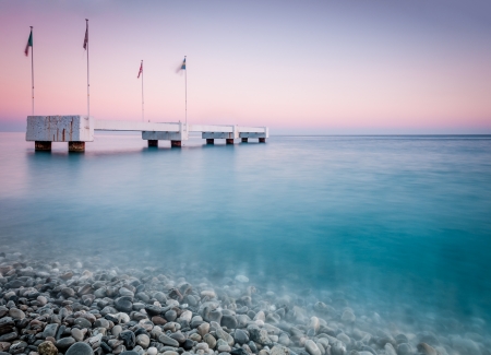 Sunset or sunrise scene with the old pier in the calm sea. Shore with pebbles in foreground and sky in background (Nice, France, Europe).の写真素材