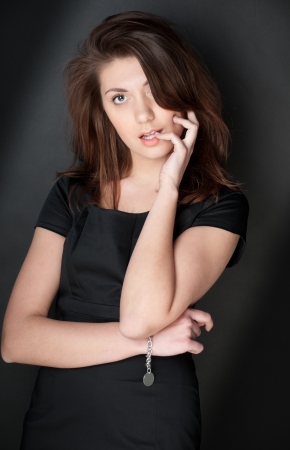 portrait of beautiful and sexy young woman in black dress with dark hair. Looking into the camera, hand at her face, dark background.の写真素材