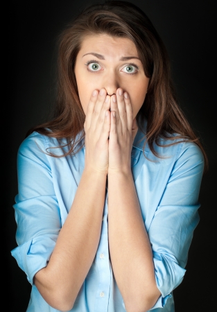 Beautiful and shocked young woman. Problems or bad news. Hands at her face. Looking into the camera, dark background.の写真素材