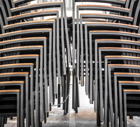 Group of black chairs stacked together in two high rows standing on grey tiled floor. Extra furniture stored in pile outdoor. Closed street cafe in morning.の写真素材