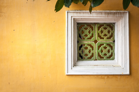 White wooden window with ornate green framing on wall of yellow stucco. Some green leaves over old worn window. House exterior in Vietnam, architecture of Asia.の写真素材
