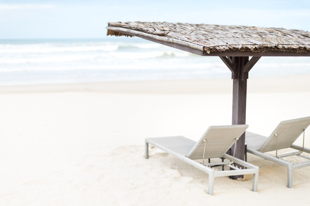 Two empty chaise longues under wooden shed on beach with blue ocean waves in background. Heaven and paradise atmosphere. Summer holidays and vacations in tropical countries.の写真素材