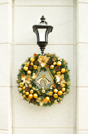 Beautiful Christmas wreath with golden balls, bells and pine cones hanging on lantern on white stone wall in street. Traditional festive decorations and design. Christmas and New Year holidays.の写真素材