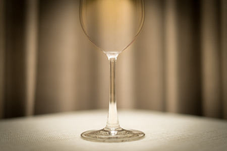 Close-up of empty wineglass on table with white tablecloth. Shiny single glass. Blurred brown drapery in background.の写真素材