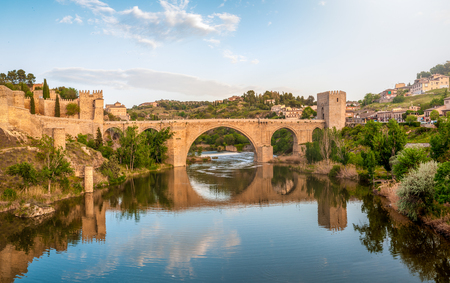Beautiful landscape of Toledo in Spain. Stone bridge across calm river. Blue sky reflected in crystal clear water. Big fort and country houses in background. Popular tourist place in Europe.の写真素材