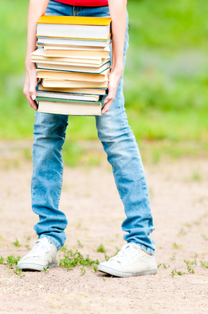 young student girl holding big pile of heavy books, lower body part. Summer or spring green park in backgroundの写真素材
