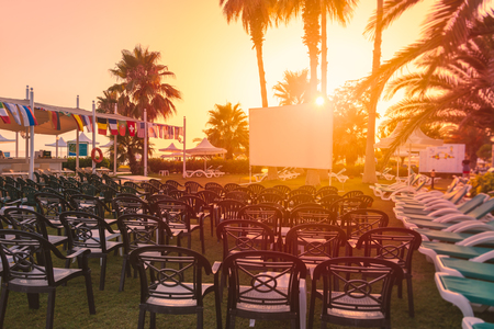 White cinema screen for projector in front of rows of chairs. Tropical scene at hotel resort at sunset.のeditorial素材