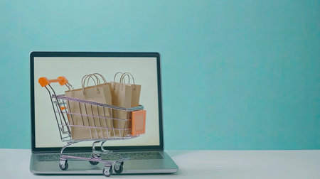 Online shopping concept with a blank notebook screen and a small shopping cart loaded with brown paper bags, highlighting e-commerce in a modern, abstract way.の素材