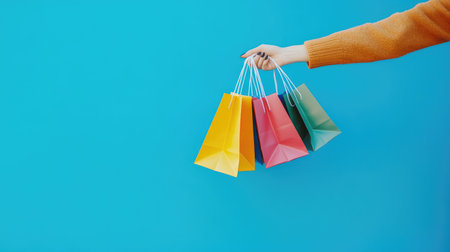 Stylish female hand holding colorful shopping bags against a vibrant blue background, with ample room for promotional messaging.の素材