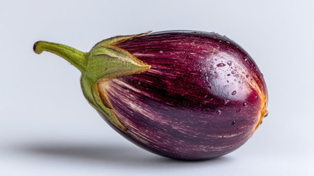 A vibrant purple eggplant sat on a clean white surface, glistening with droplets of water, perfect for showcasing the beauty of fresh produce in culinary designs.の素材