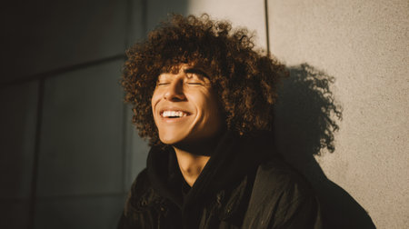 A joyful young man with curly hair smiles brightly while basking in natural light, creating a warm and positive urban portrait.の素材