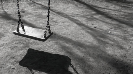 A striking black and white photograph featuring an empty swing casting a shadow on bare ground, capturing themes of solitude and nostalgia.の素材