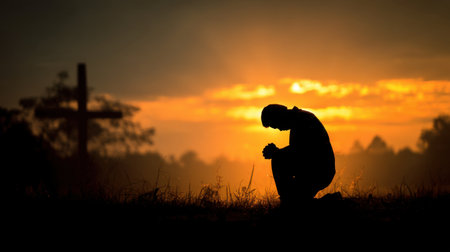 A serene image depicting a person kneeling in prayer during a stunning sunset, with a cross silhouette creating a profound atmosphere of spirituality and reflection.の素材