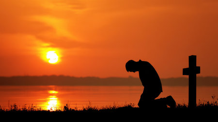 A silhouette captures a person kneeling in prayer against a stunning sunset, with a cross nearby, evoking feelings of peace and reflection.の素材