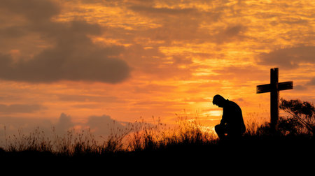 A serene silhouette of a person praying against a beautiful sunset, featuring a cross in the background, symbolizing faith and reflection.の素材