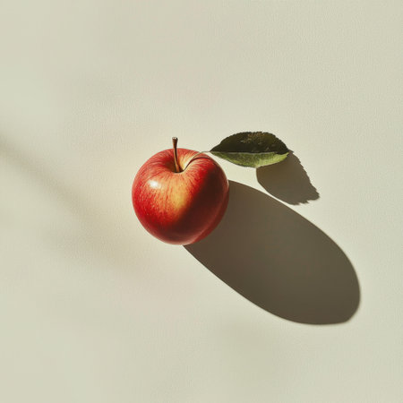 A vibrant red apple rests on a white surface, accompanied by a green leaf. The soft natural light creates gentle shadows, enhancing its fresh look.の素材