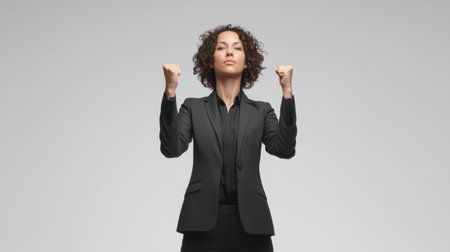 A confident businesswoman in a stylish suit stands with raised fists, showcasing empowerment and determination against a neutral backdrop, embodying strength and motivation.の素材