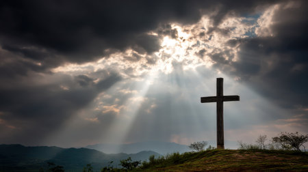 A stunning silhouette of a cross stands atop a hill, framed by dramatic clouds and radiant beams of sunlight in a serene mountainous setting.の素材