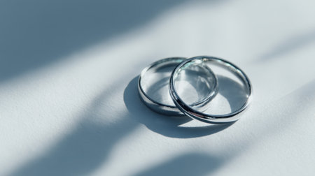 Captivating close-up of two elegant silver wedding rings resting on a white surface, softly illuminated to emphasize their beauty and simplicity.の素材