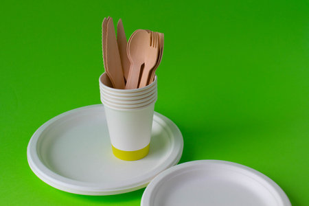 Eco-friendly disposable dishes made of bamboo wood and paper on a green background. Draped spoons, forks, knives, bamboo bowls with paper cupsの写真素材