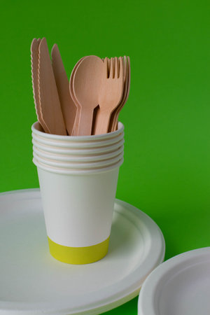 Eco-friendly disposable dishes made of bamboo wood and paper on a green background. Draped spoons,...
