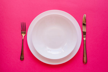 The concept of gastronomy and food. Top view of an empty white plate with fork and knife on a pink background. Serving dinner in a restaurant.の写真素材