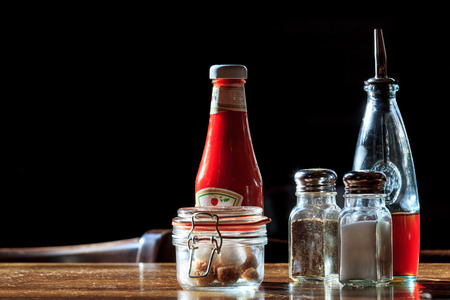 Typical pub utensils like ketchup, sugar, salt, pepper and vinegal on a wooden table on black...