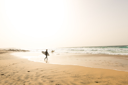 Two male surfers in their wetsuits walking to the ocean waves on an empty beach with their surfboards at sunsetの写真素材