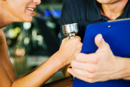 Woman with dumbbells in a gym, her personal trainer gives a report on her training (close-up, not faces to be seen, focus on the dumbbells)の写真素材