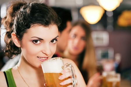 Group of three friends in a bar drinking beer - selective focus on beautiful woman in front zipping from her glassの写真素材