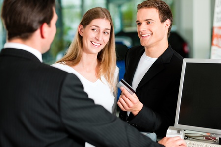 Sales situation in a car dealership, the young couple is giving the credit card to pay for the new car Sales situation in a car dealership, the young couple is giving the credit card to pay for the new car