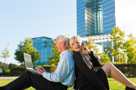 Business people working outdoors on a meadow - he is working with laptop, she is calling someone on phoneの写真素材