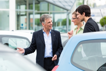 Mature car dealer and young couple standing on parking place at dealership in front of carsの写真素材