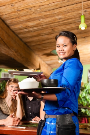 Young people eating in a Thai restaurant; the waitress brings the dishes, rice and othersの写真素材