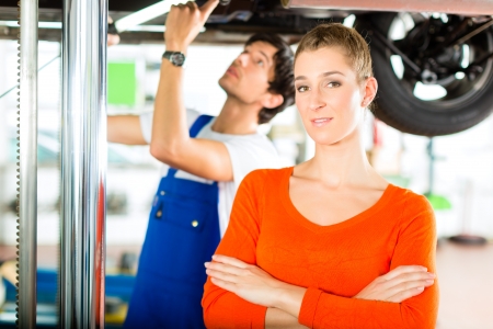 Woman standing in front of her car which is lifted on an auto hoist, a mechanic doing a repair underneath the auto の写真素材